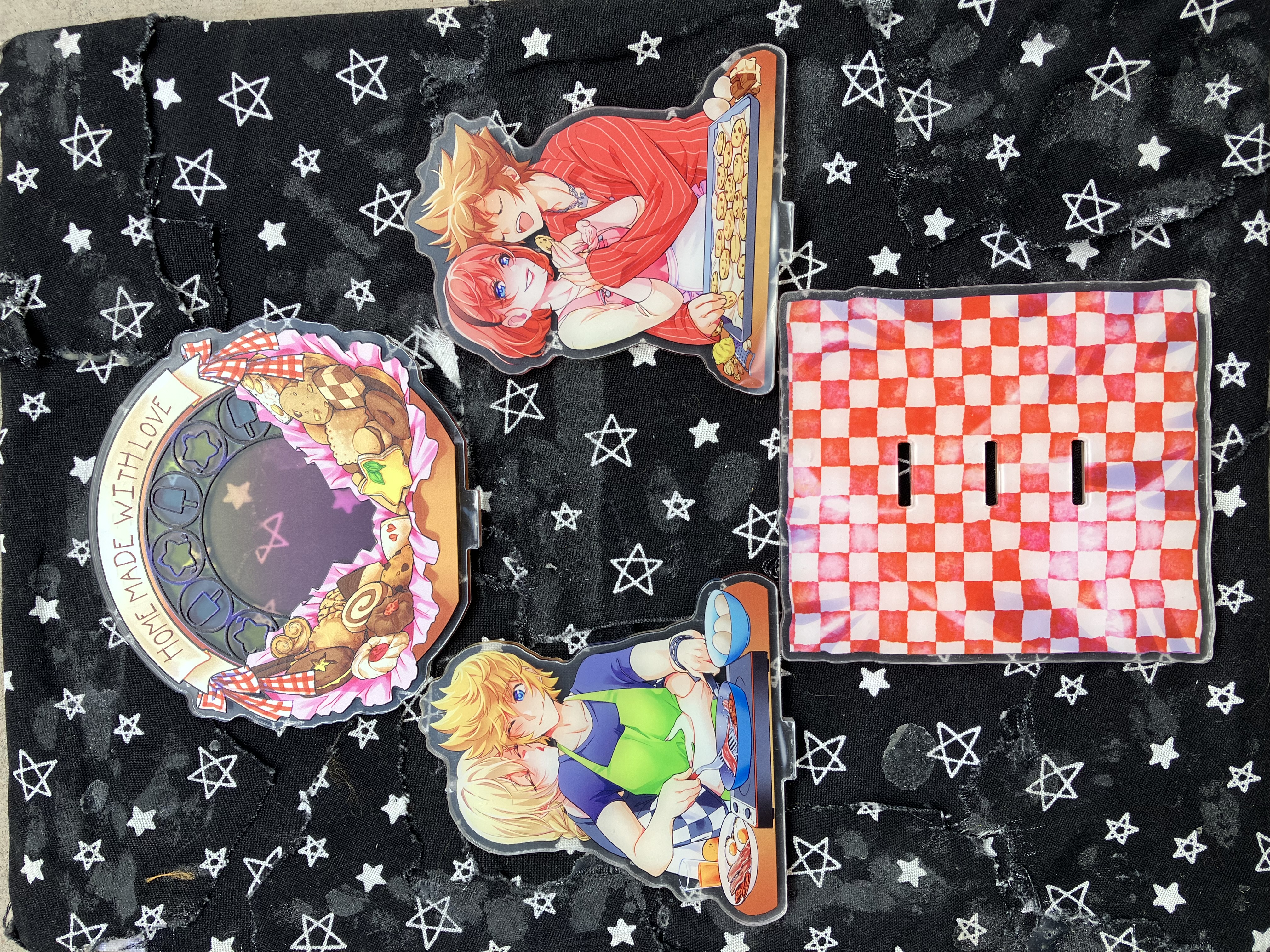 Front of acrylic standee of Kingdom hearts Kairi, Roxas, Namine, and Sora in casual clothes with food surrounding them in a kitchen. Roxas is cooking bacon and eggs while Namine kisses his cheek. Kairi is feeding cookies to Sora